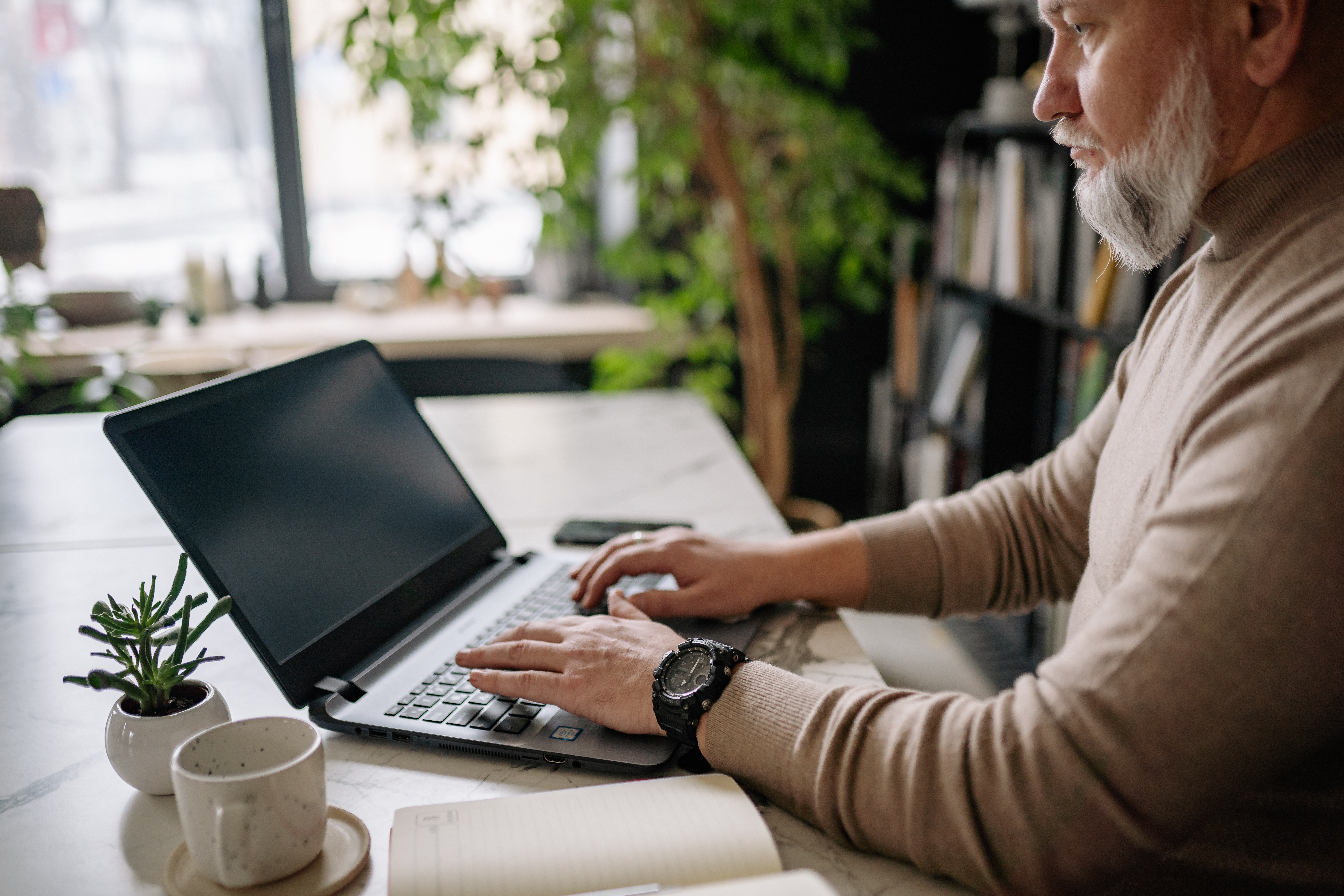 Homem branco de barca branca, usando moletom bege e relógio preto, sentado à mesa mexendo no notebook.
